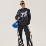 Person wearing a black tracksuit with '75' logo and sunglasses, holding a blue bag on a white background