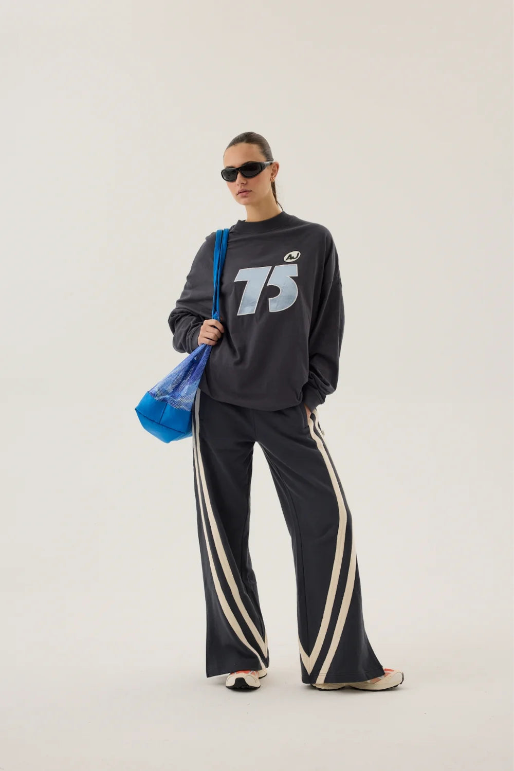 Person wearing a black tracksuit with '75' logo and sunglasses, holding a blue bag on a white background