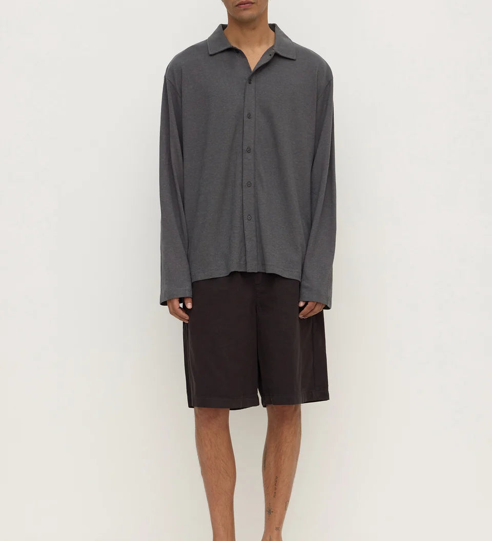 A man modeling a dark gray long-sleeve button-up shirt and dark brown shorts, paired with black flip-flop sandals, against a plain off-white studio background.