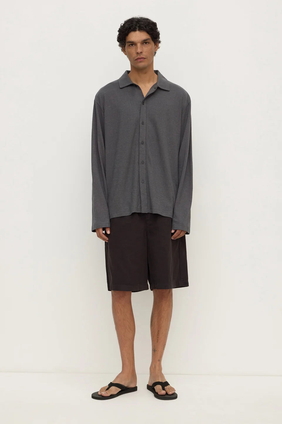 A man modeling a dark gray long-sleeve button-up shirt and dark brown shorts, paired with black flip-flop sandals, against a plain off-white studio background.