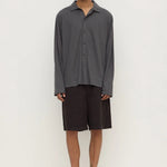 A man modeling a dark gray long-sleeve button-up shirt and dark brown shorts, paired with black flip-flop sandals, against a plain off-white studio background.