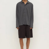 A man modeling a dark gray long-sleeve button-up shirt and dark brown shorts, paired with black flip-flop sandals, against a plain off-white studio background.