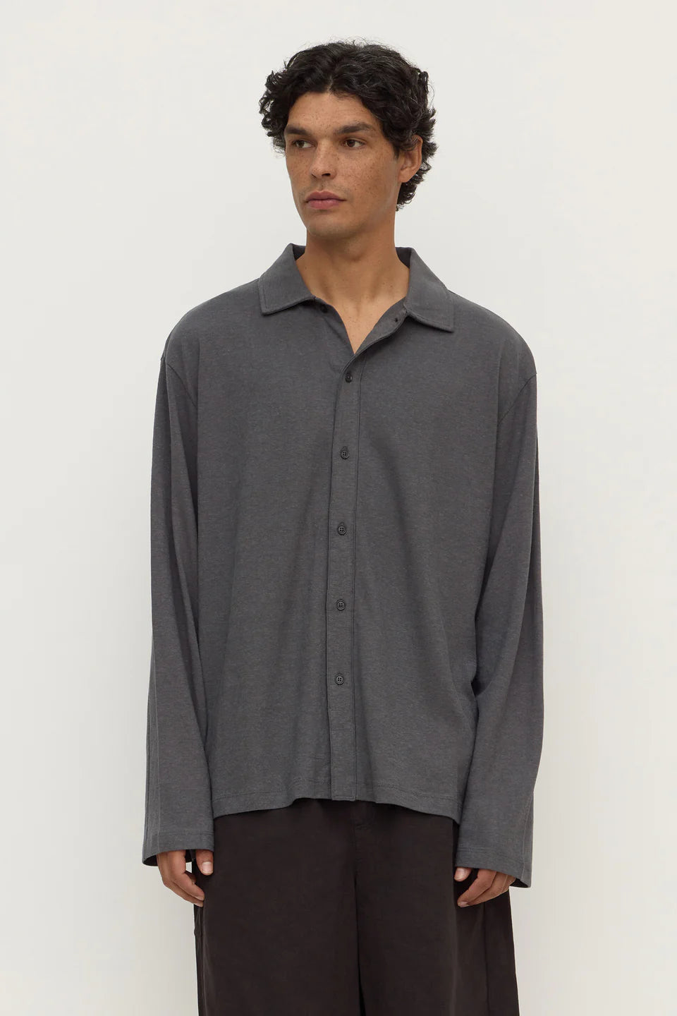A man wearing a dark gray long-sleeve button-up shirt with a relaxed, oversized fit and dark brown trousers, standing against a plain off-white background.