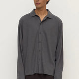 A man wearing a dark gray long-sleeve button-up shirt with a relaxed, oversized fit and dark brown trousers, standing against a plain off-white background.