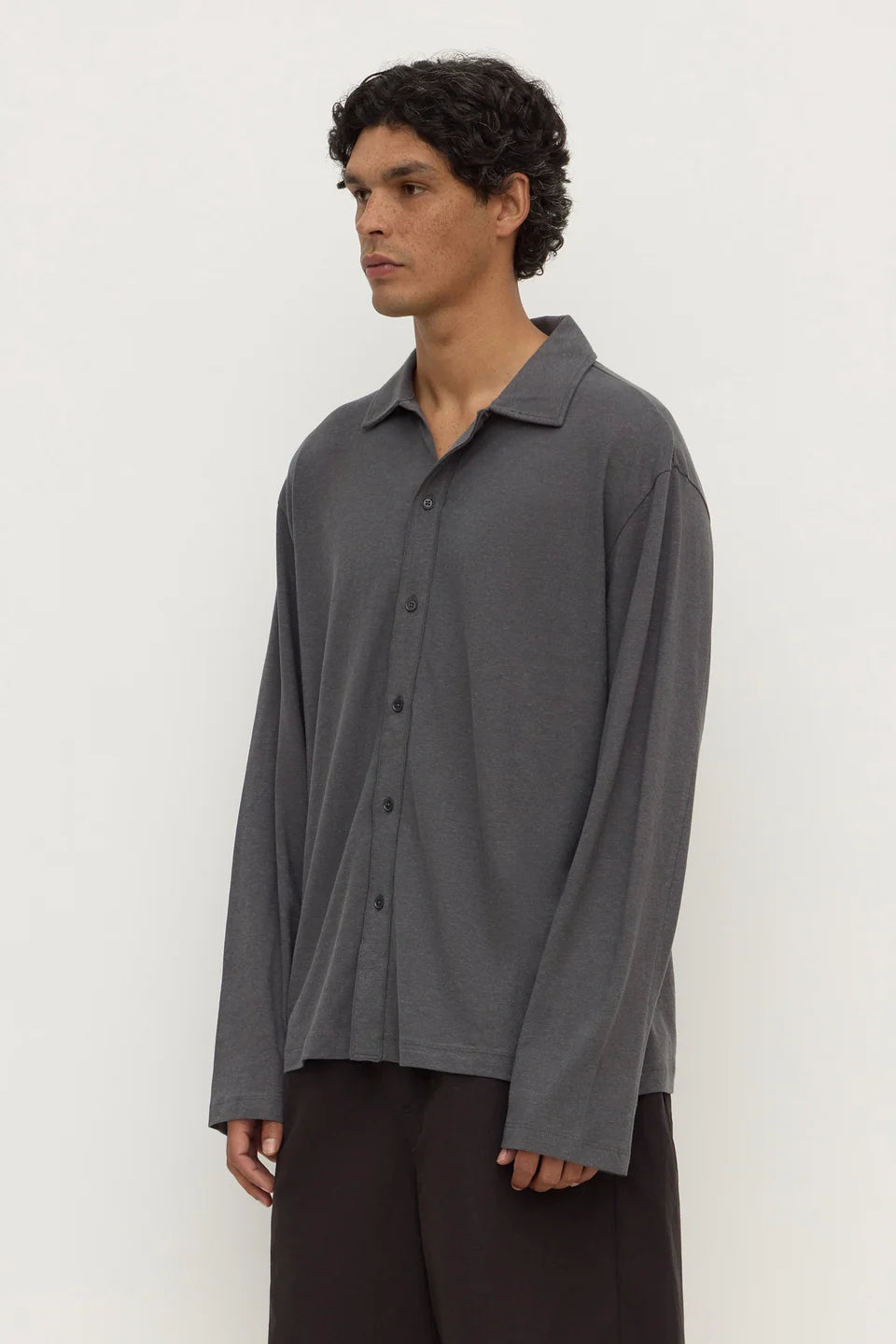 A man wearing a dark gray long-sleeve button-up shirt and dark brown pants, standing against a plain off-white background.
