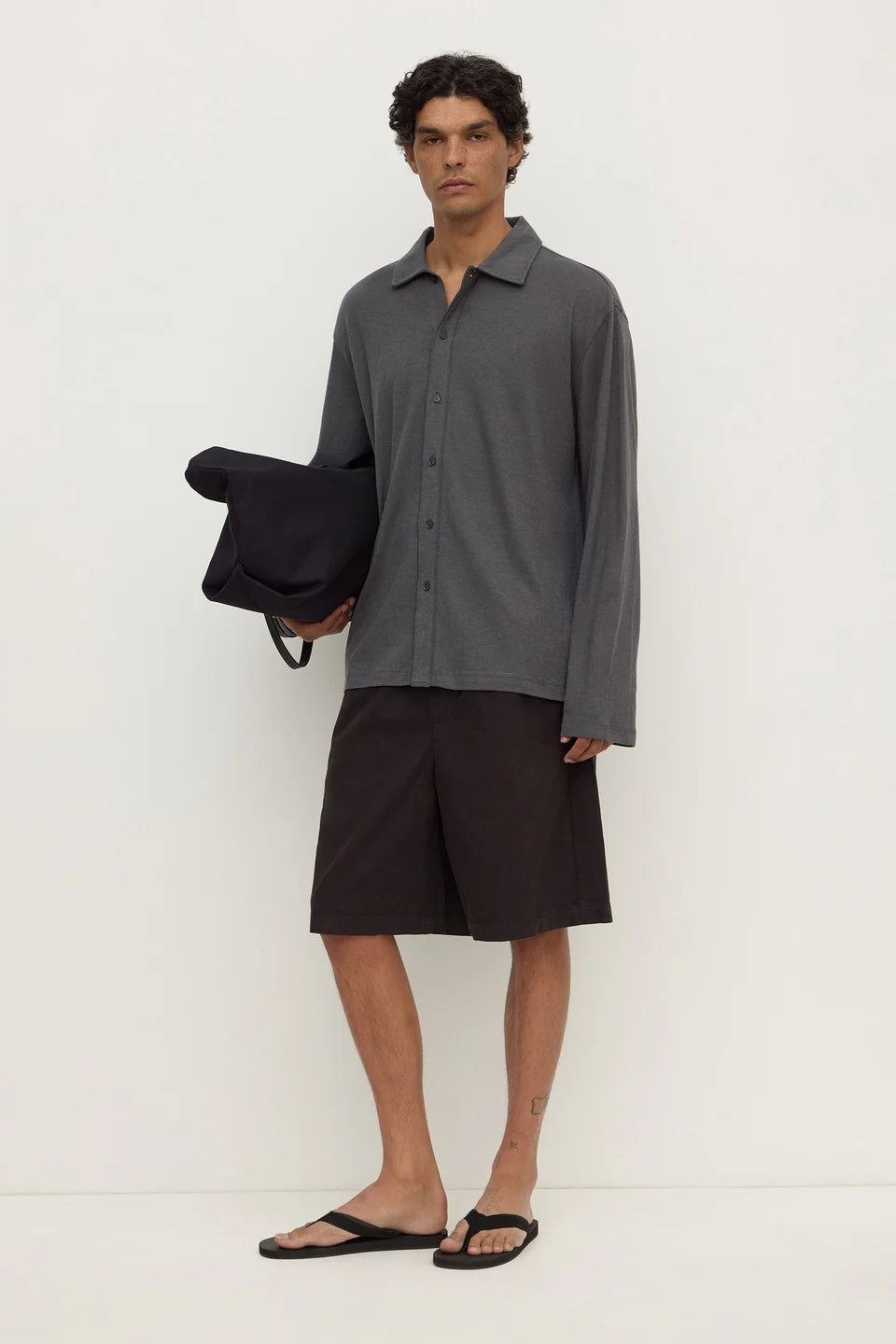 A male model stands against a plain off-white studio wall wearing a dark gray long-sleeve button-up shirt, dark brown shorts, and black flip-flops, while holding a black bag.