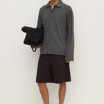 A male model stands against a plain off-white studio wall wearing a dark gray long-sleeve button-up shirt, dark brown shorts, and black flip-flops, while holding a black bag.