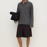 A male model stands against a plain off-white studio wall wearing a dark gray long-sleeve button-up shirt, dark brown shorts, and black flip-flops, while holding a black bag.