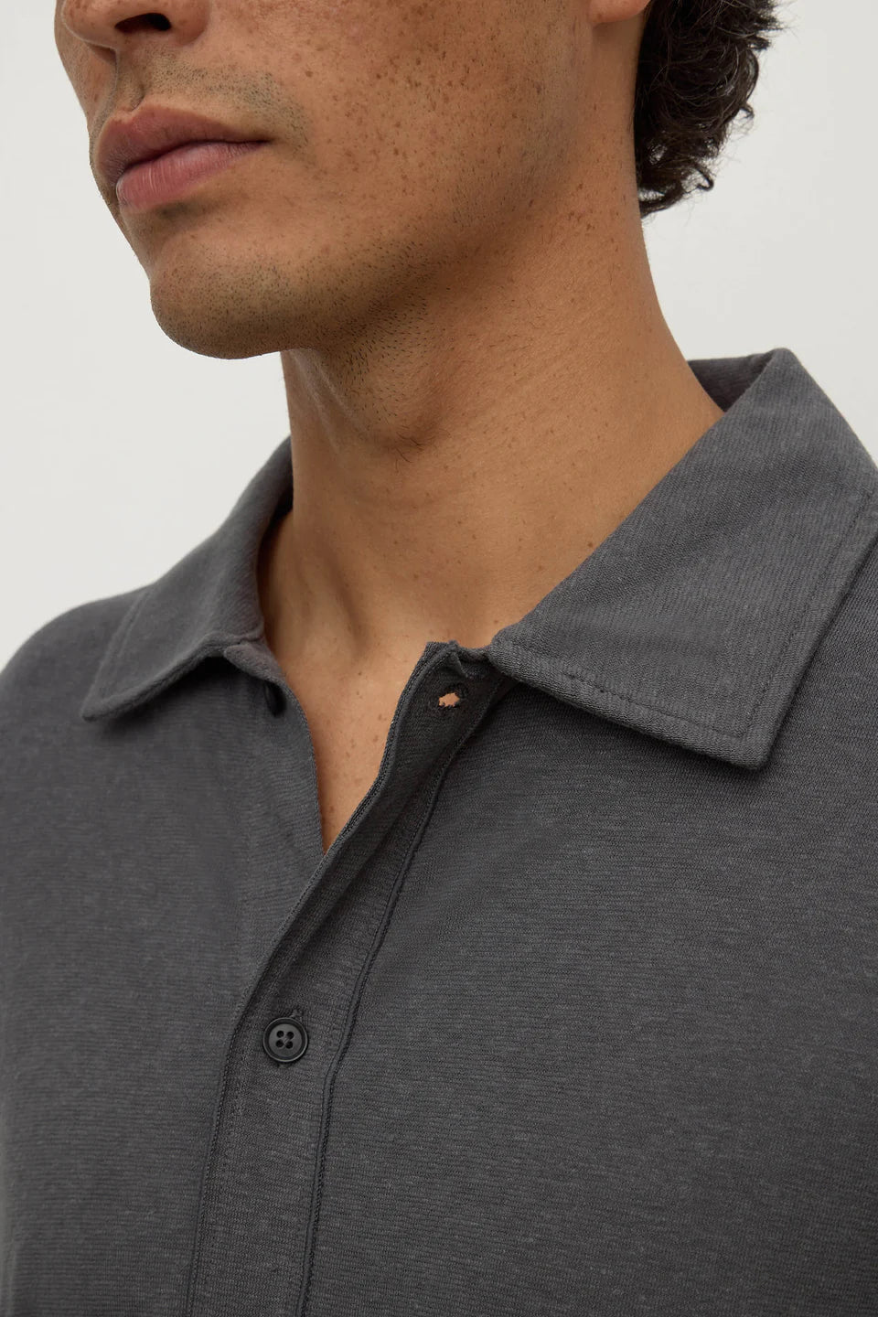 Close-up of a man wearing a dark gray heathered polo shirt, showing the collar, button placket, and part of his neck and lower face against a plain light background.