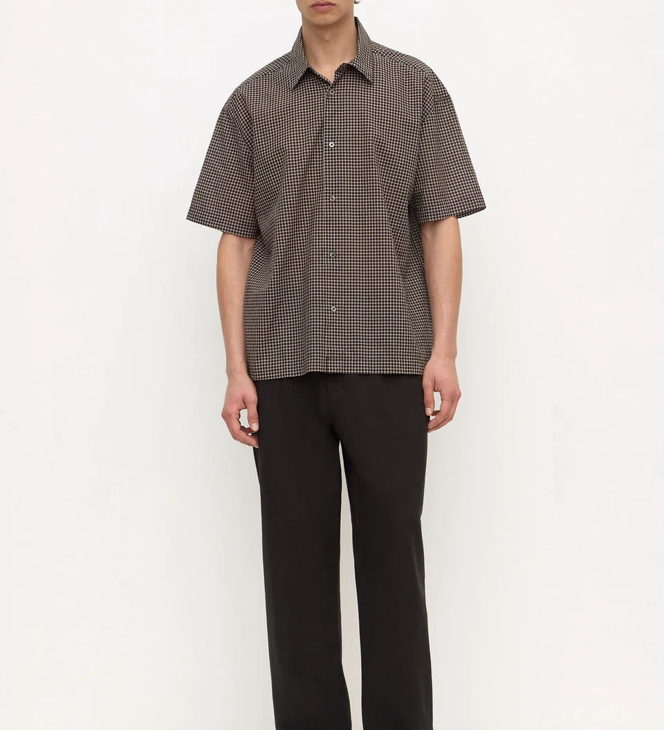 A man stands against a plain white studio background wearing a short-sleeve button-up shirt with a fine brown and black checkered pattern, paired with black trousers and black shoes.
