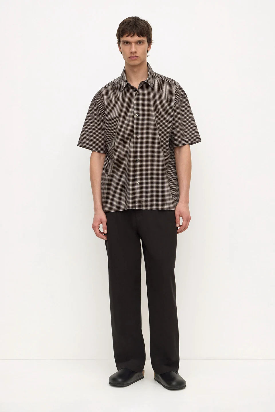 A man stands against a plain white studio background wearing a short-sleeve button-up shirt with a fine brown and black checkered pattern, paired with black trousers and black shoes.