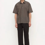 A man stands against a plain white studio background wearing a short-sleeve button-up shirt with a fine brown and black checkered pattern, paired with black trousers and black shoes.