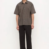 A man stands against a plain white studio background wearing a short-sleeve button-up shirt with a fine brown and black checkered pattern, paired with black trousers and black shoes.