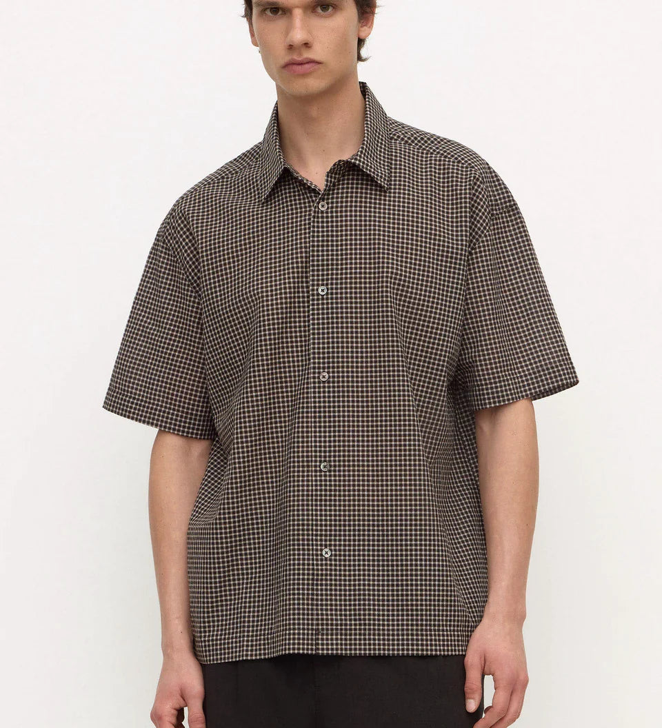 A man wearing a short-sleeve button-up shirt with a brown and black micro-check pattern, paired with dark trousers, standing against a light gray studio background.