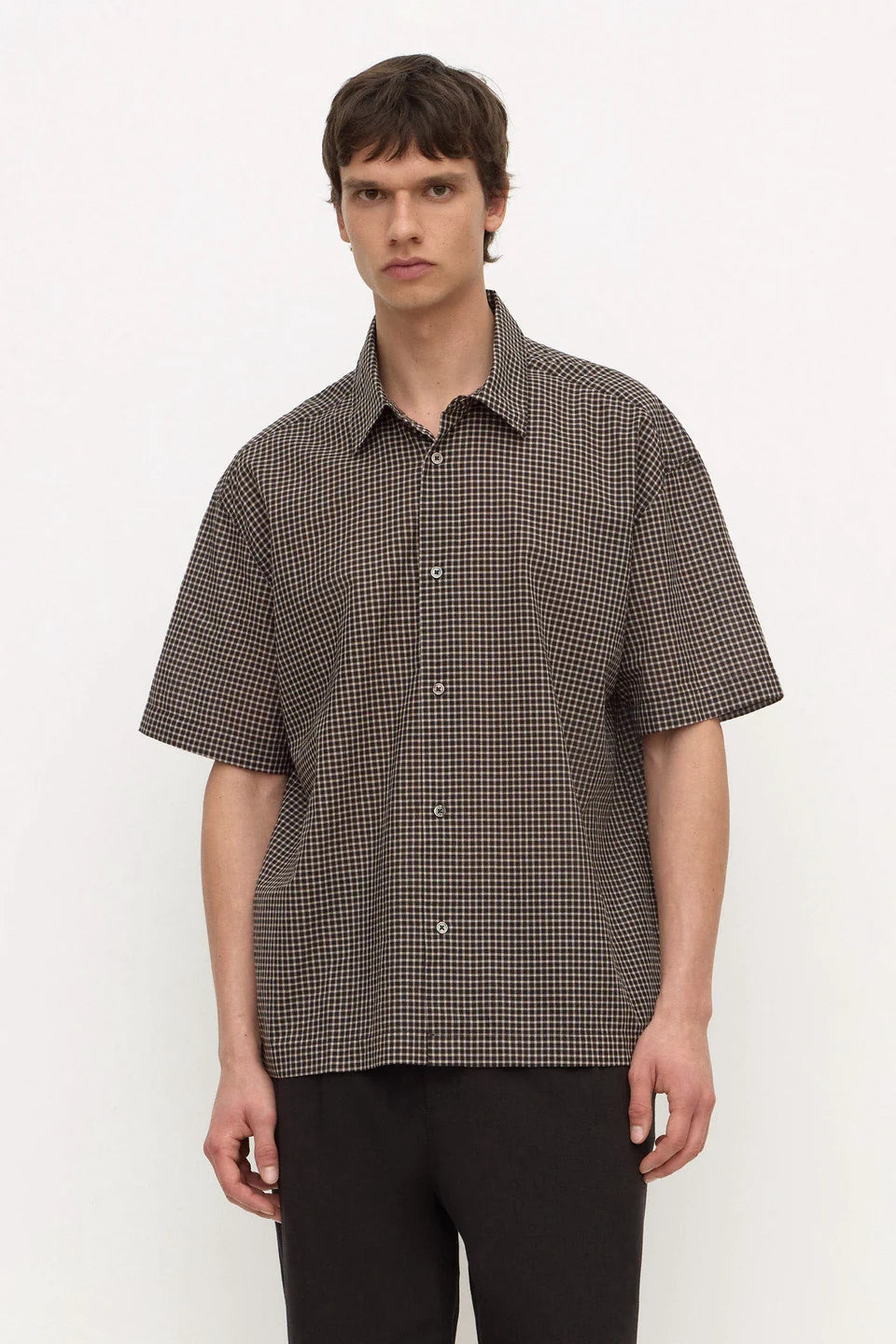 A man wearing a short-sleeve button-up shirt with a brown and black micro-check pattern, paired with dark trousers, standing against a light gray studio background.