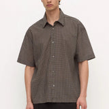 A man wearing a short-sleeve button-up shirt with a brown and black micro-check pattern, paired with dark trousers, standing against a light gray studio background.