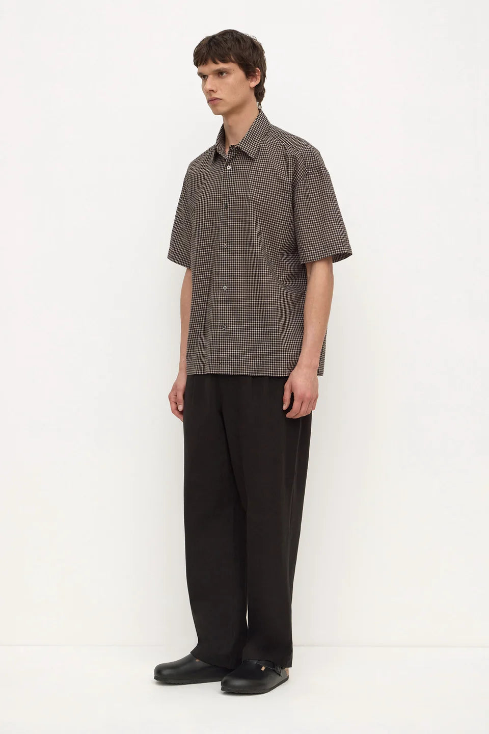 A man modeling a short-sleeve button-up shirt with a fine brown and gray checkered pattern, paired with loose black trousers and black shoes, standing against a plain white studio background.