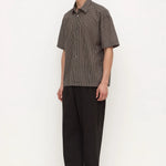 A man modeling a short-sleeve button-up shirt with a fine brown and gray checkered pattern, paired with loose black trousers and black shoes, standing against a plain white studio background.