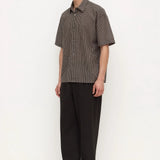 A man modeling a short-sleeve button-up shirt with a fine brown and gray checkered pattern, paired with loose black trousers and black shoes, standing against a plain white studio background.