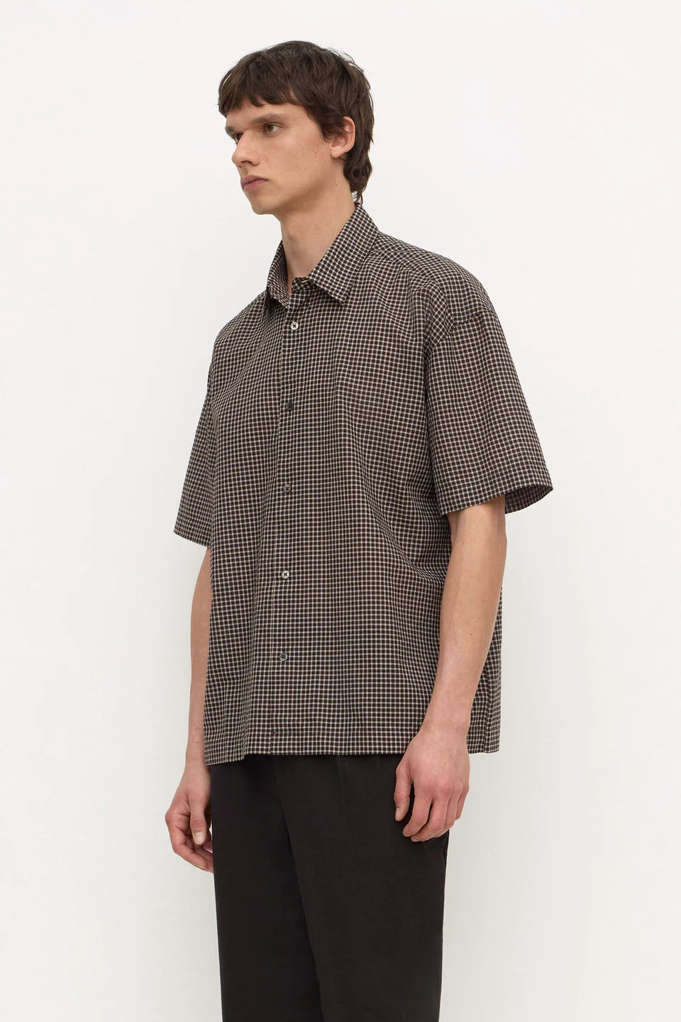 A man wearing a short-sleeve button-up shirt with a brown and black micro-check pattern, paired with black trousers, standing against a plain white studio background.