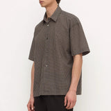 A man wearing a short-sleeve button-up shirt with a brown and black micro-check pattern, paired with black trousers, standing against a plain white studio background.