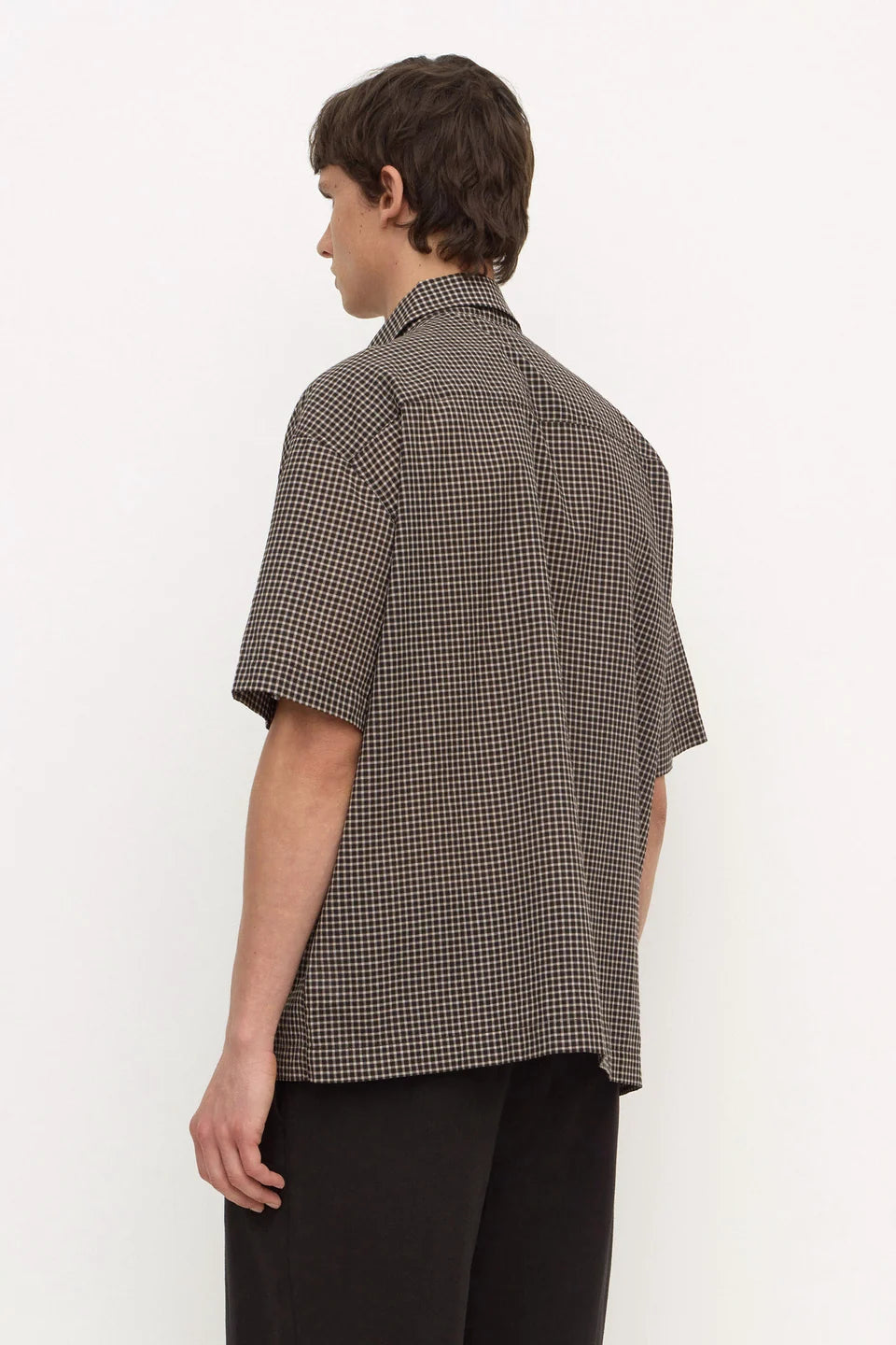A man shown from the back wearing a short-sleeve button-up shirt with a small-scale brown and white micro-check pattern, paired with black trousers, against a plain white studio background.