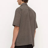 A man shown from the back wearing a short-sleeve button-up shirt with a small-scale brown and white micro-check pattern, paired with black trousers, against a plain white studio background.