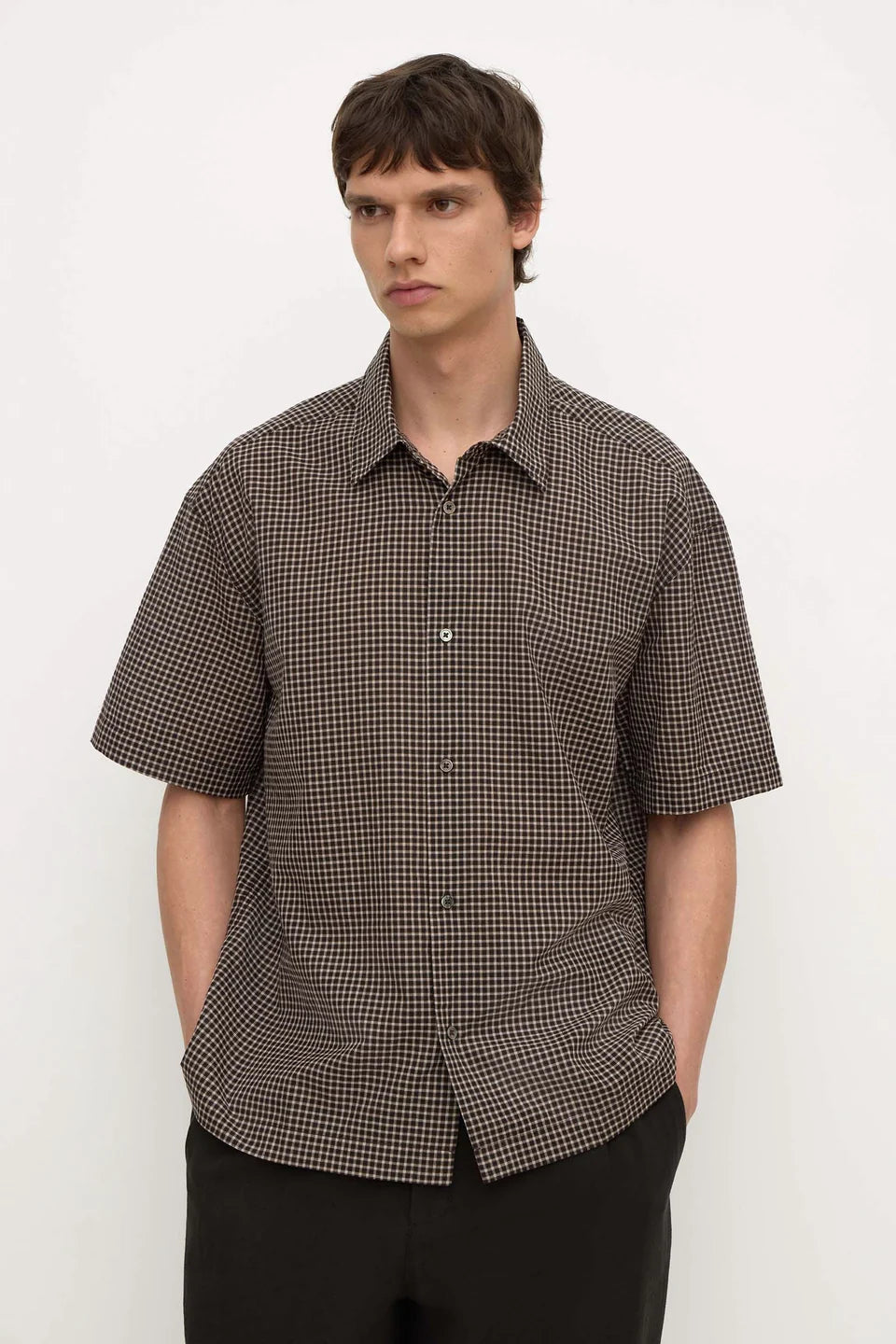 A man wearing a short-sleeve button-up shirt with a brown and black micro-check pattern, paired with black trousers, standing against a plain white background.