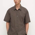 A man wearing a short-sleeve button-up shirt with a brown and black micro-check pattern, paired with black trousers, standing against a plain white background.