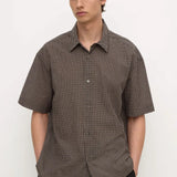 A man wearing a short-sleeve button-up shirt with a brown and black micro-check pattern, paired with black trousers, standing against a plain white background.