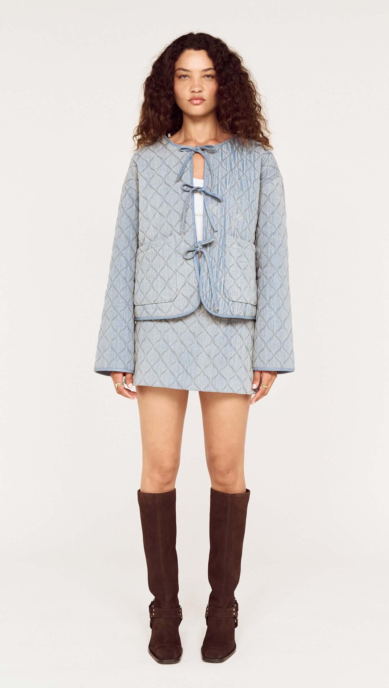 Woman wearing a light blue textured jacket and skirt set with brown knee-high boots on a white background