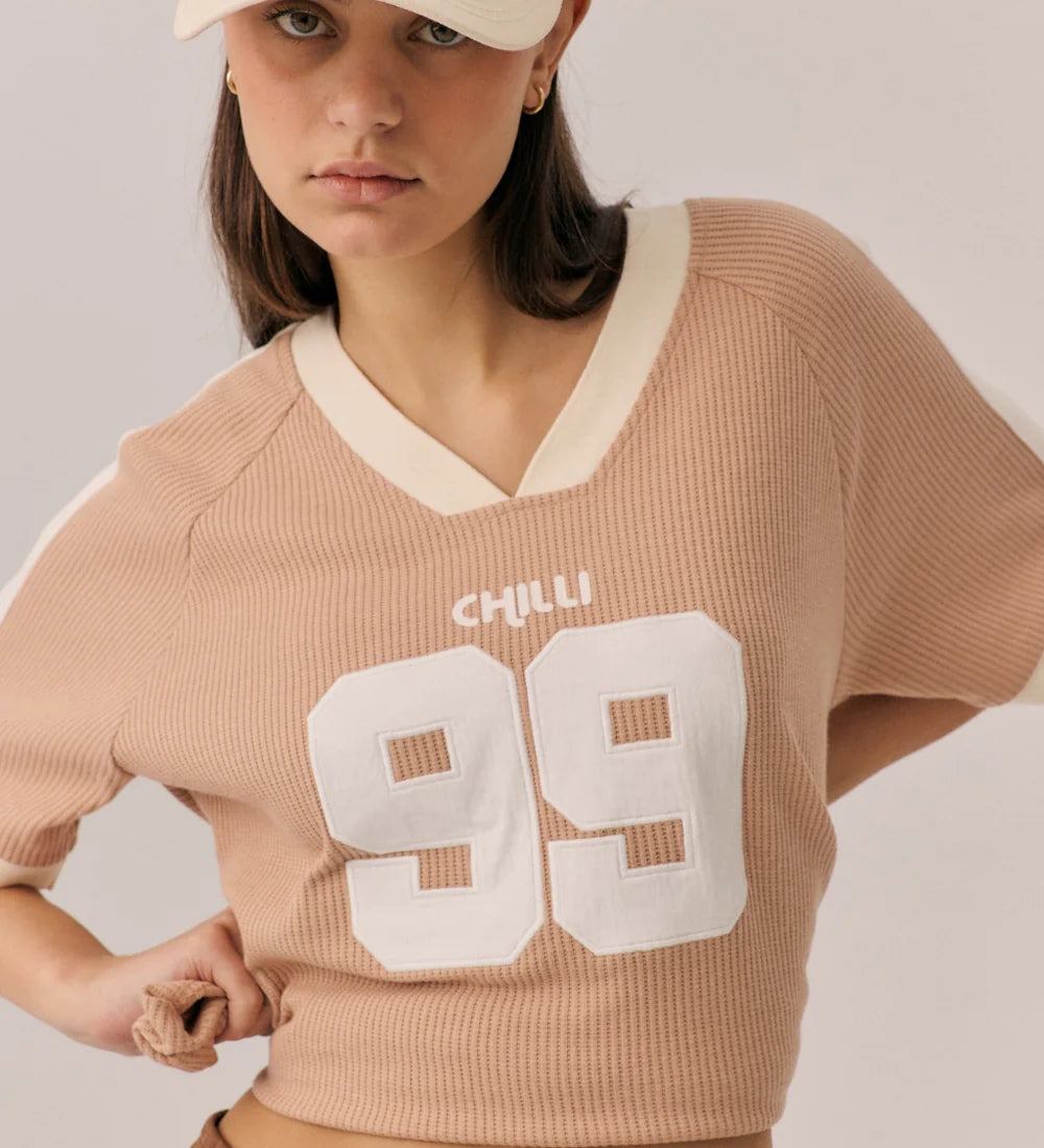 Woman wearing a beige sweater with 'CHILLI 99' text, brown pants, and a beige cap with red text on a plain background.