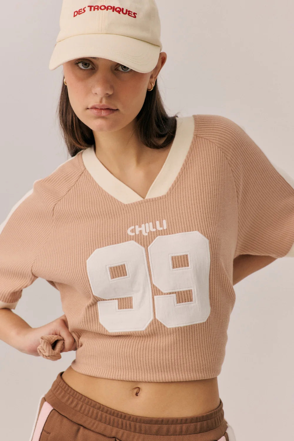 Woman wearing a beige sweater with 'CHILLI 99' text, brown pants, and a beige cap with red text on a plain background.