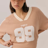 Woman wearing a beige sweater with 'CHILLI 99' text, brown pants, and a beige cap with red text on a plain background.
