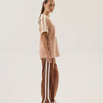 Woman wearing a beige top and brown pants with white stripes on a plain background