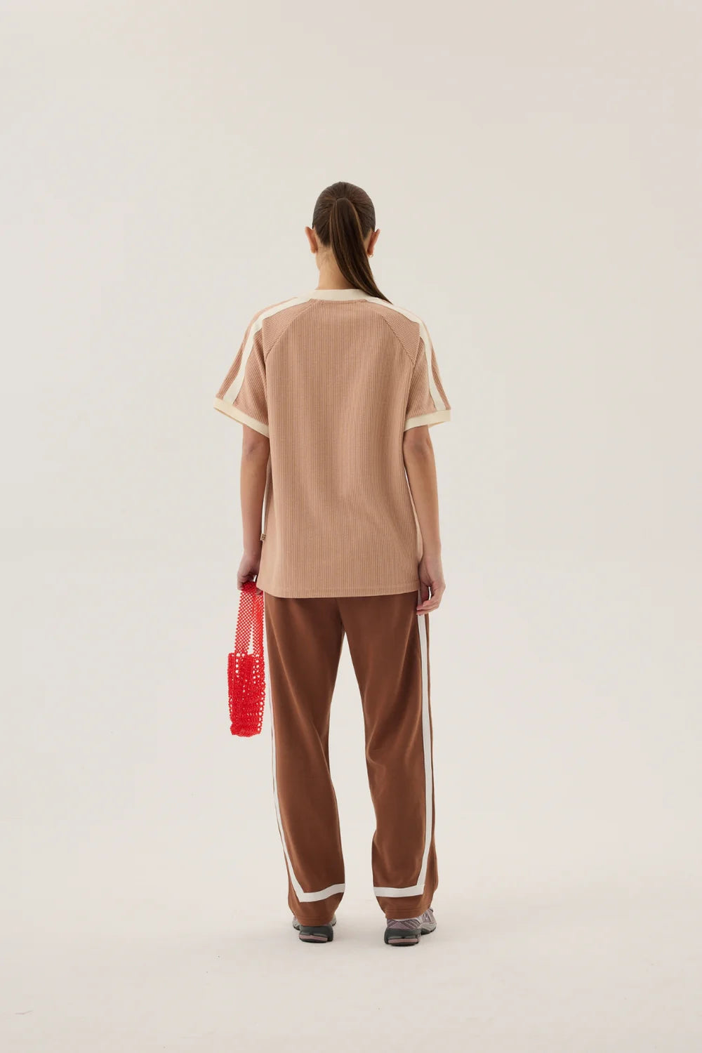 Person wearing a beige top and brown pants with white stripes, holding a red bag on a plain background