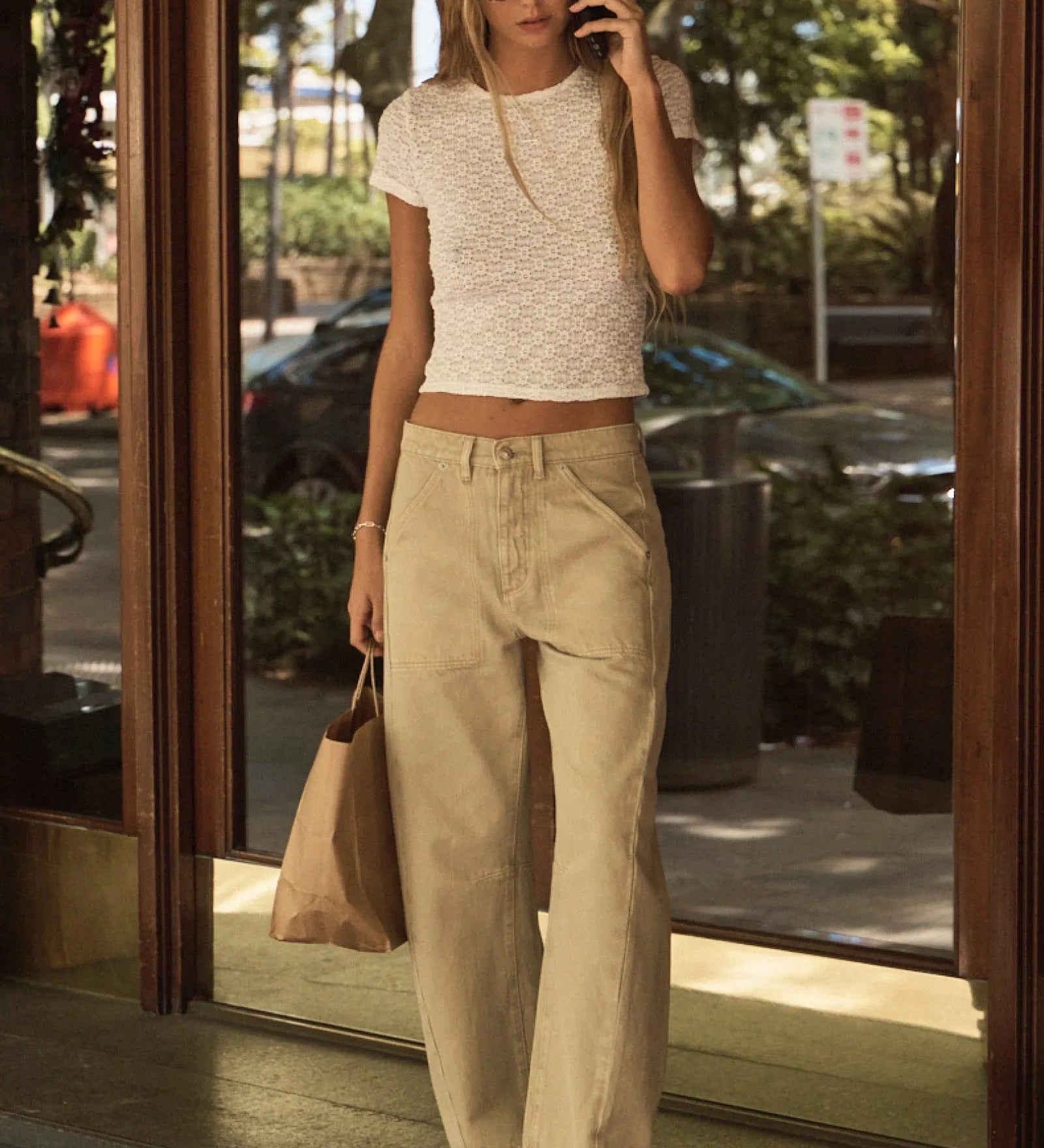 Woman in a white top and beige pants standing outside a building, talking on a phone.