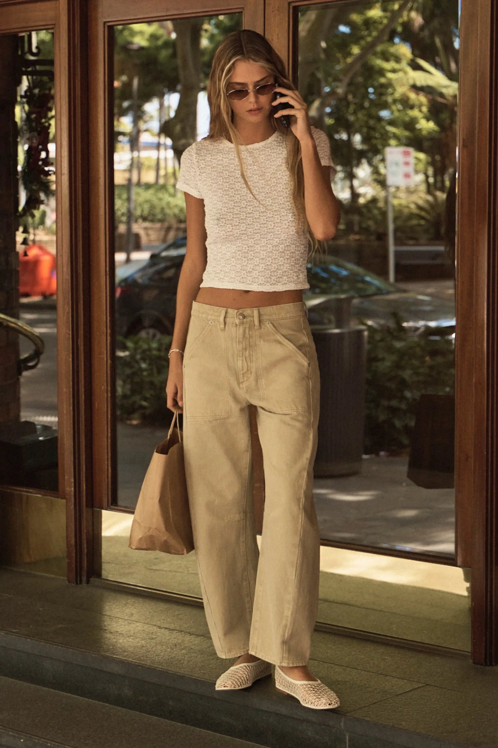 Woman in a white top and beige pants standing outside a building, talking on a phone.