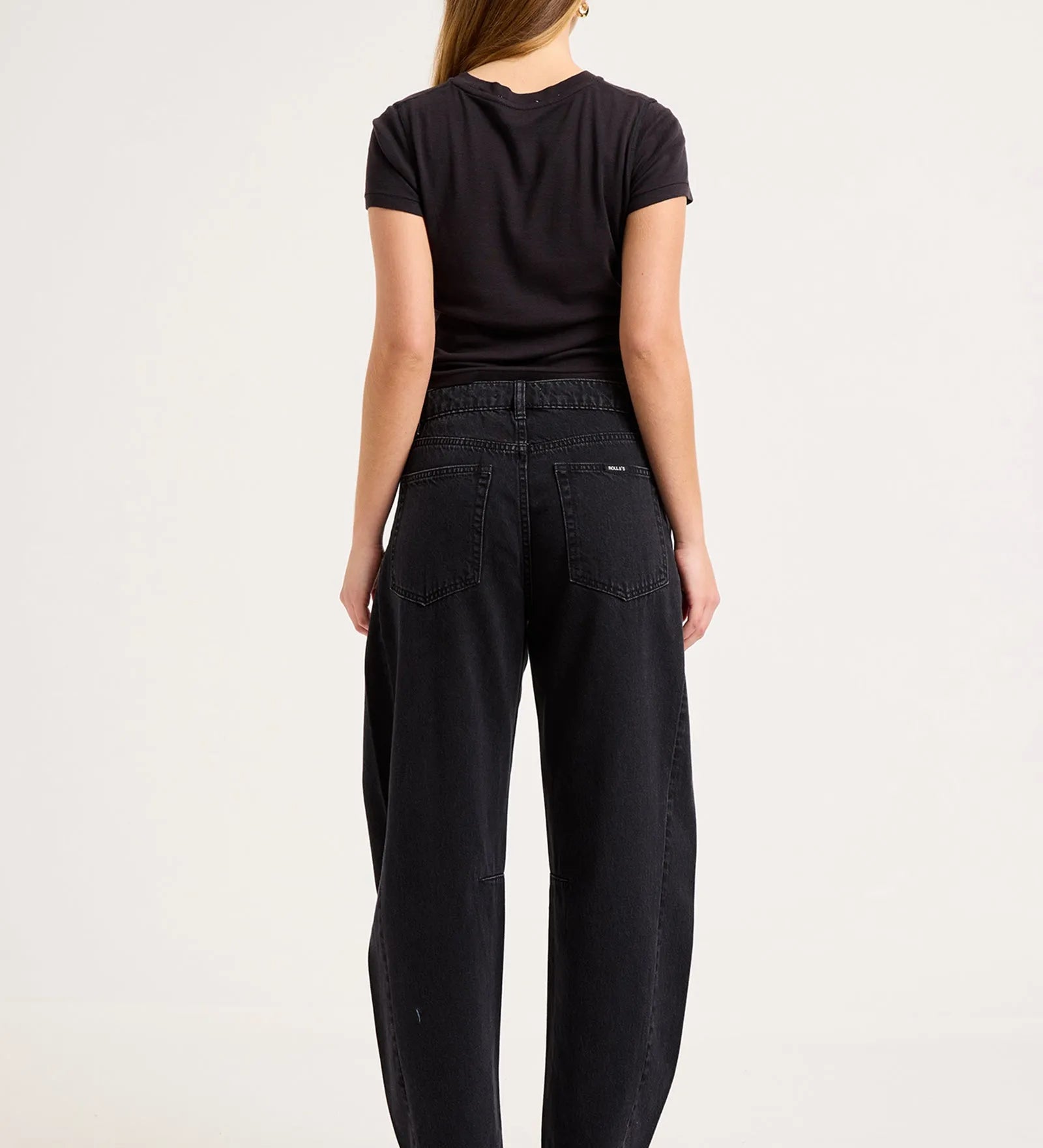 A woman viewed from the back wearing a black short-sleeve t-shirt and black wide-leg jeans, standing against a plain white studio background.