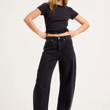 A woman modeling a black short-sleeve crop top and black wide-leg jeans with black heels, standing against a plain light gray studio background.