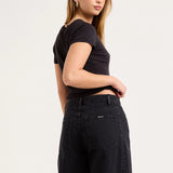 A woman wearing a black short-sleeve crop top and black wide-leg jeans, photographed from the back against a plain light gray studio background.