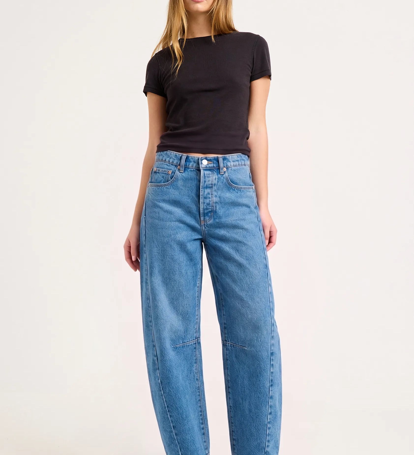 A model stands against a light gray studio background wearing a black short-sleeve crop top, wide-leg blue jeans, and black pointed heels.