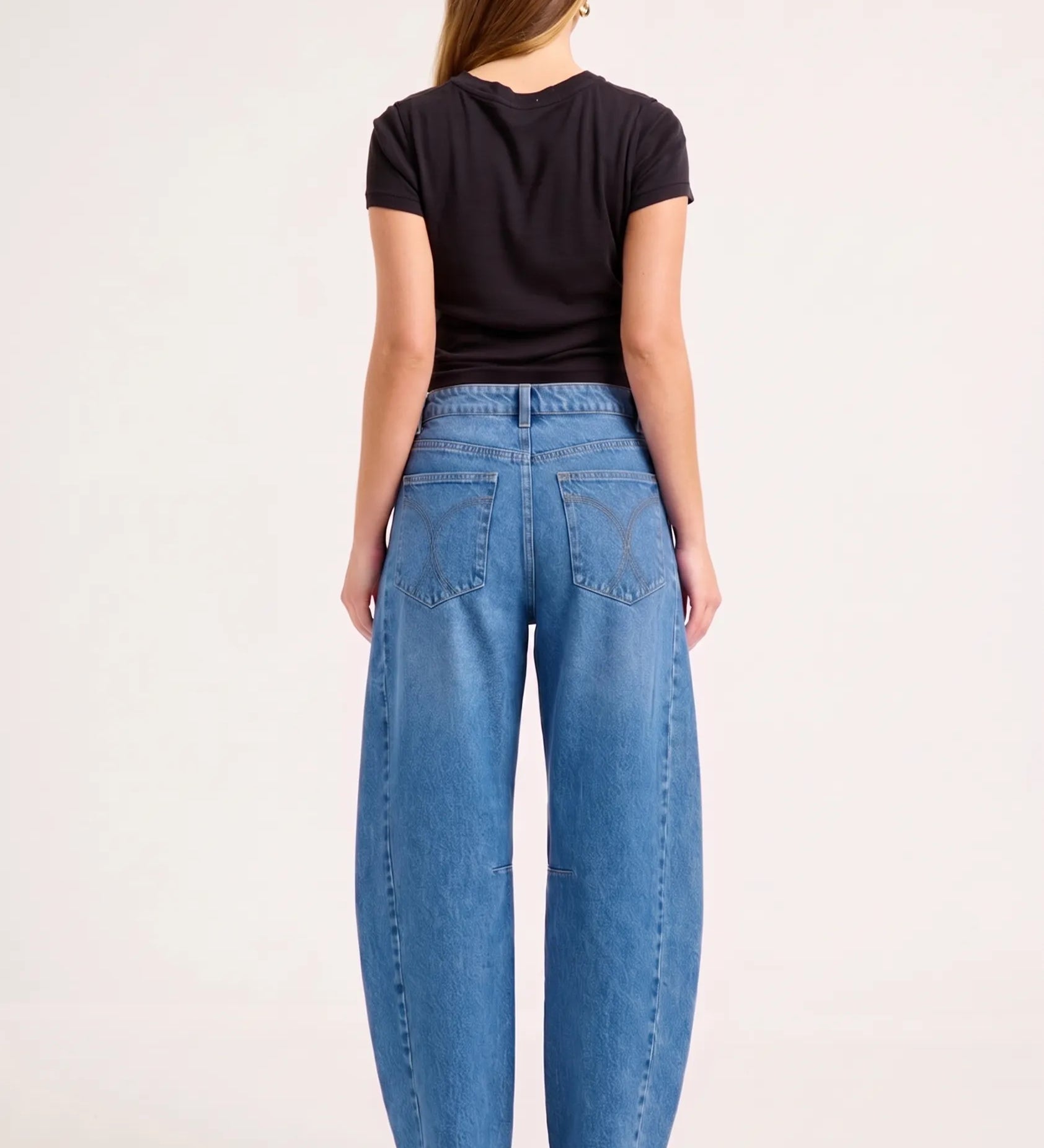 A woman viewed from the back wearing wide-leg blue jeans, a black short-sleeve top, and black heels against a plain light background.