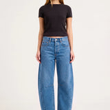 A model stands against a light gray studio background wearing a black short-sleeve crop top, wide-leg blue jeans, and black pointed heels.