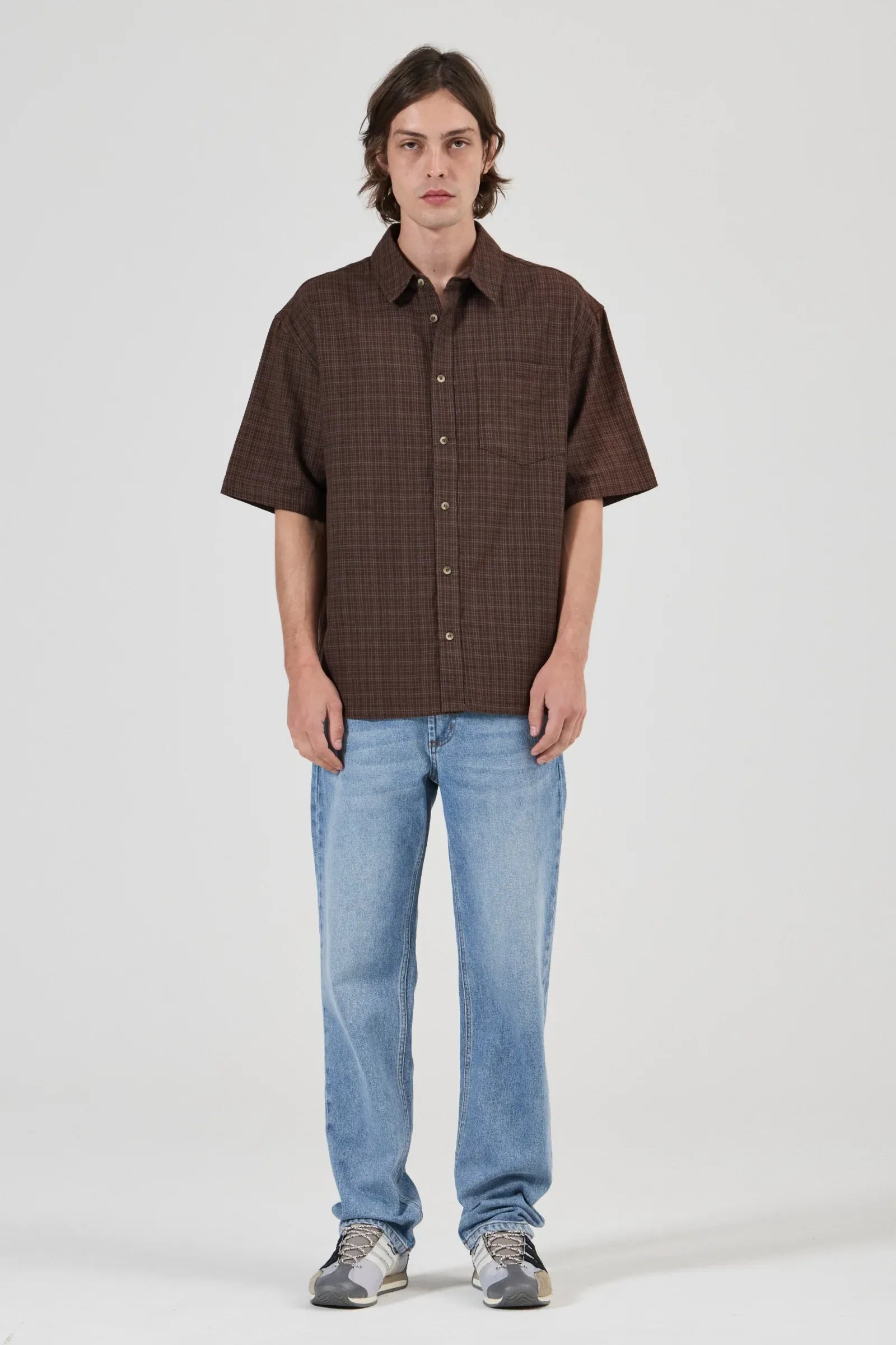 Man wearing a brown checkered shirt and light blue jeans on a white background