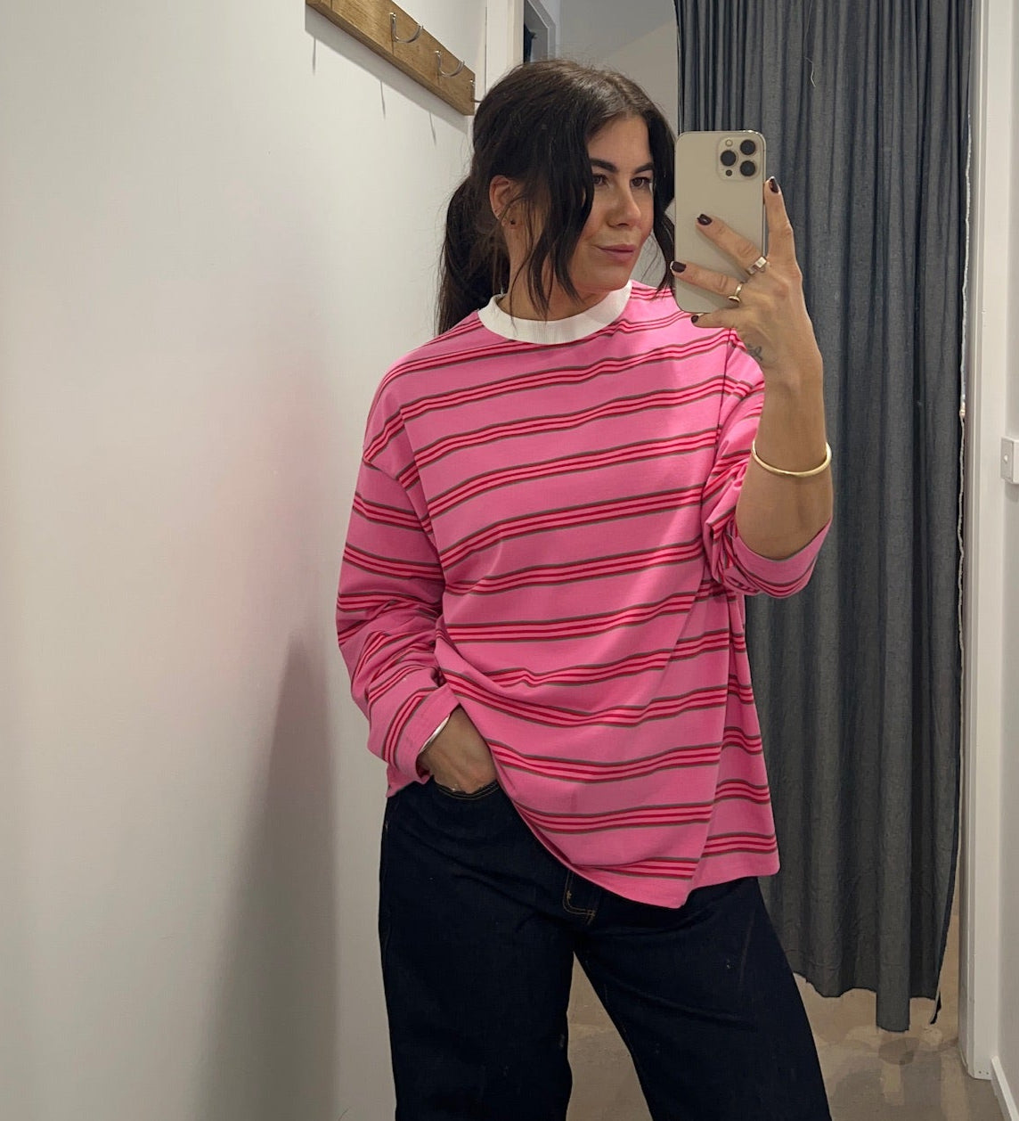 Person wearing a pink striped long sleeve  top taking a mirror selfie in a room with white walls and a gray curtain.