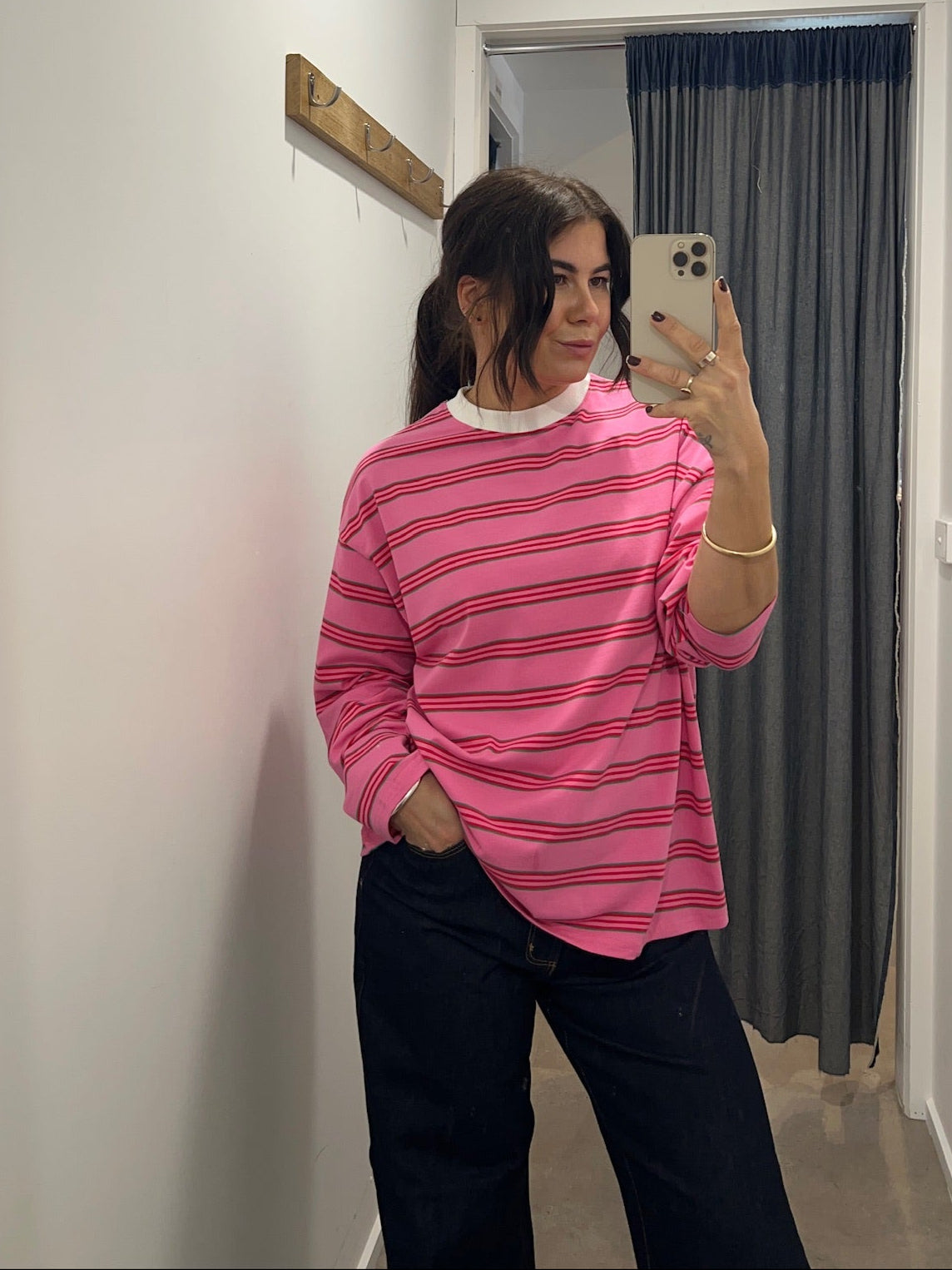 Person wearing a pink striped long sleeve  top taking a mirror selfie in a room with white walls and a gray curtain.