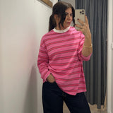 Person wearing a pink striped long sleeve  top taking a mirror selfie in a room with white walls and a gray curtain.