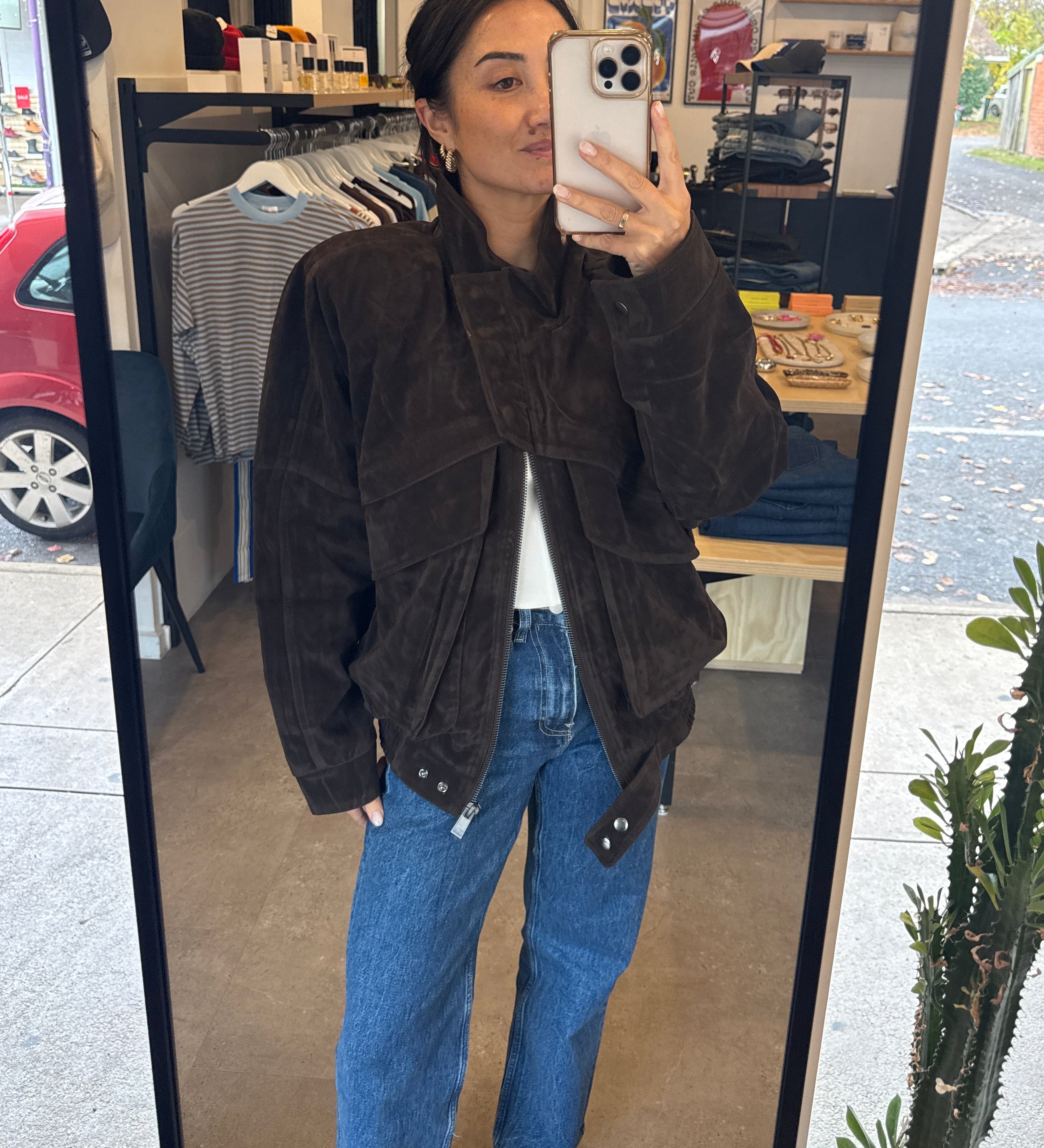 Person taking a mirror selfie wearing a brown jacket and blue jeans in an indoor setting.