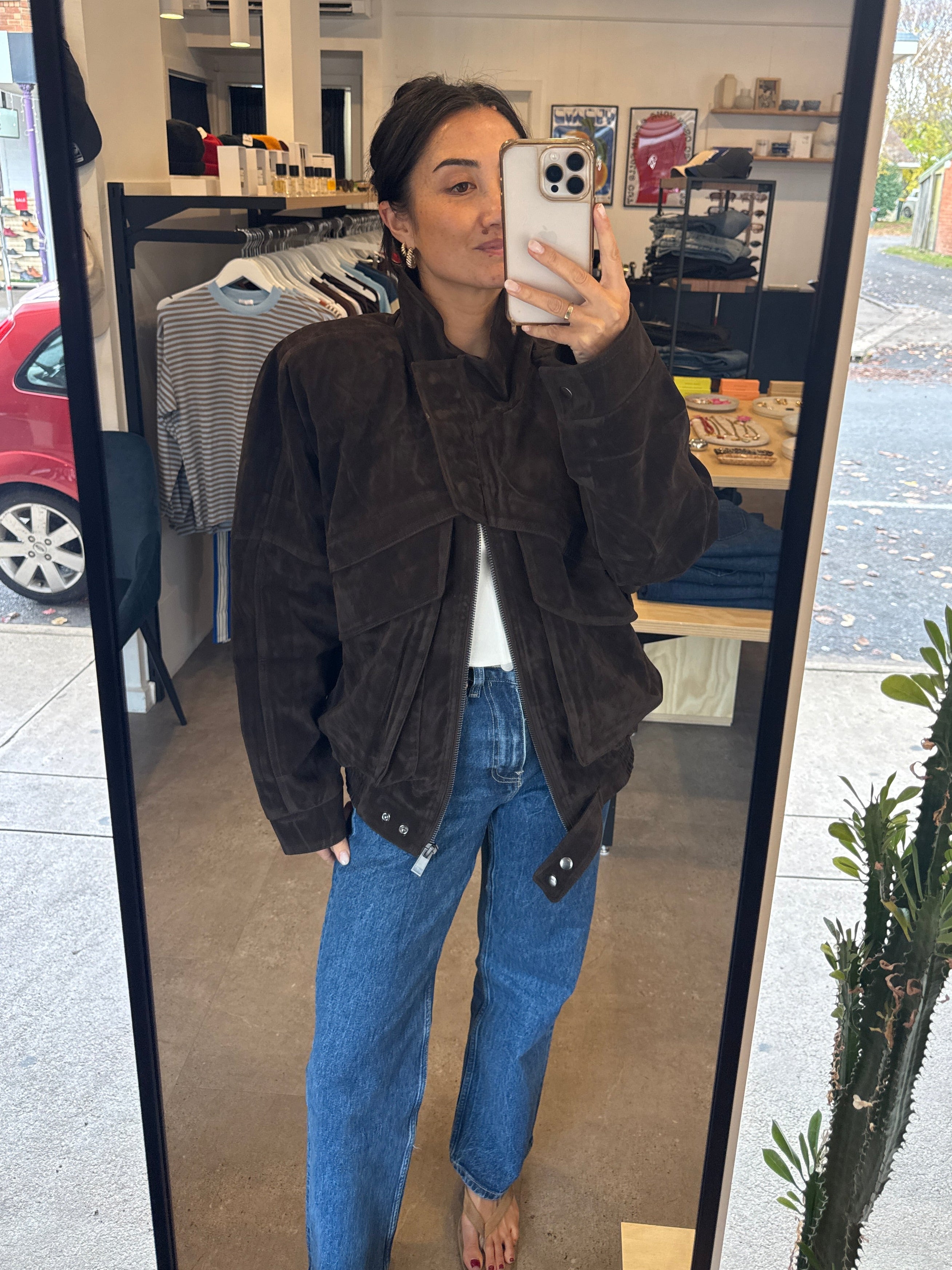 Person taking a mirror selfie wearing a brown jacket and blue jeans in an indoor setting.